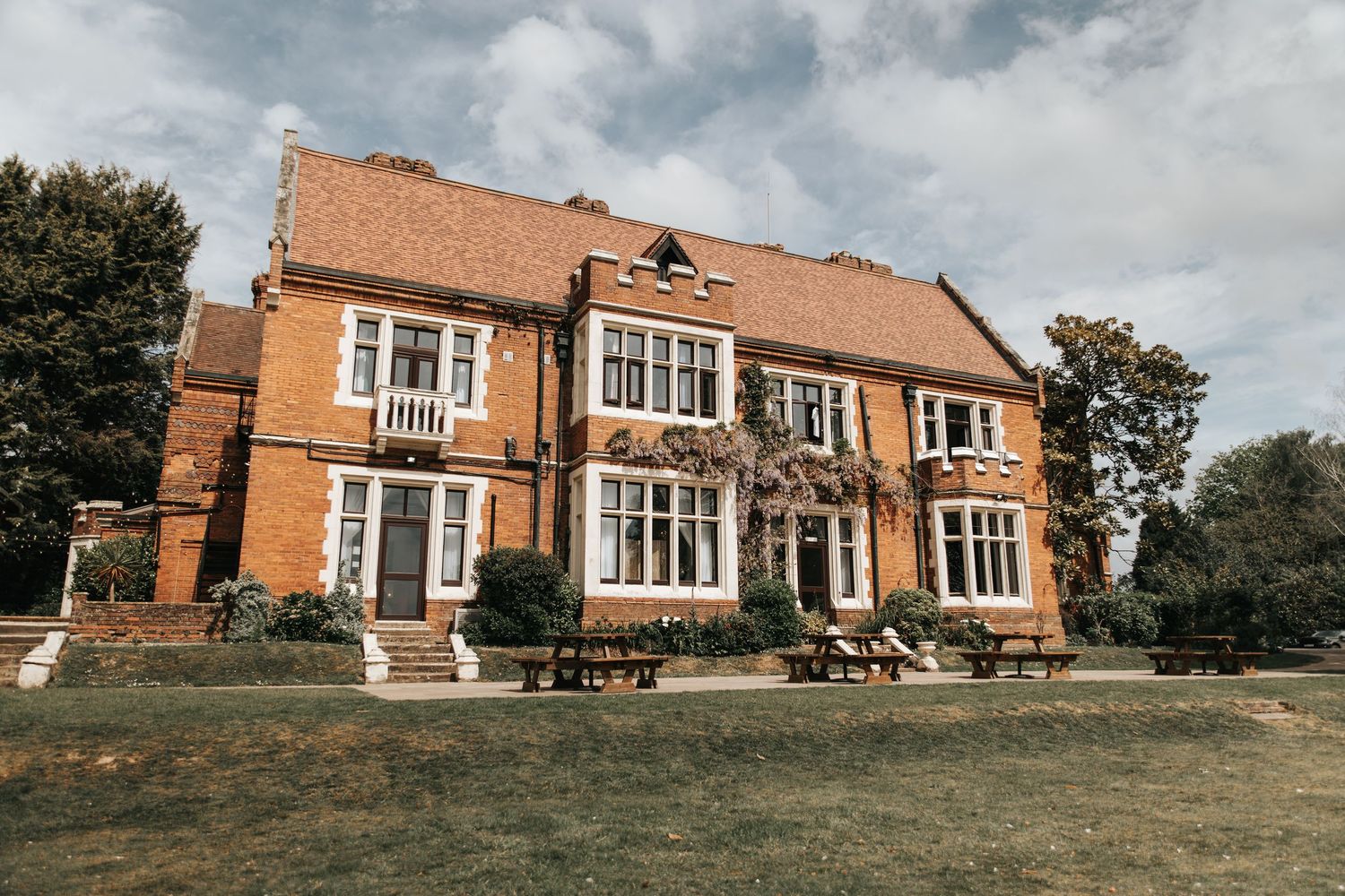 A charming red brick English country manor house with white-framed windows, surrounded by lush green lawns and picnic benches.