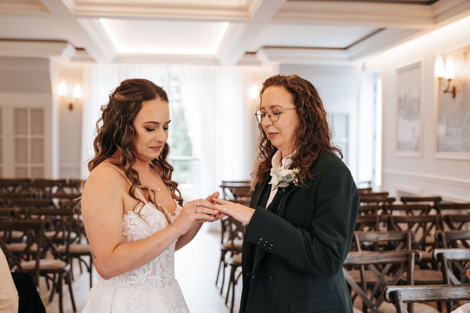 Two brides exchange rings during an elegant indoor wedding ceremony with wooden cross-back chairs in the background.