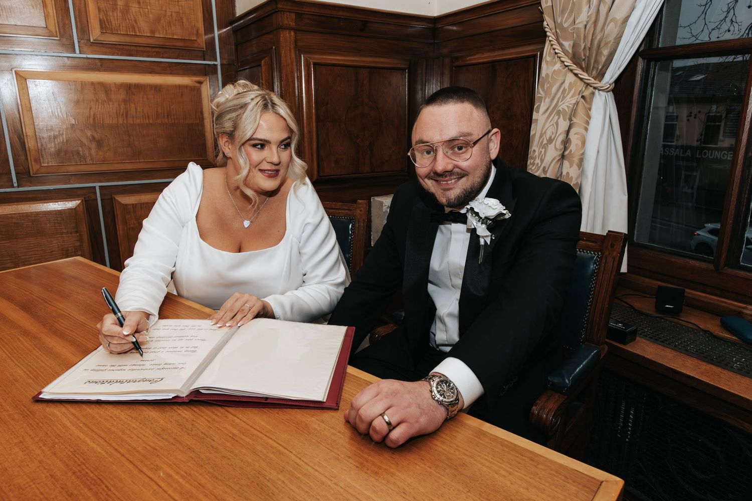 Wedding couple signs marriage certificate at wooden table in formal setting.