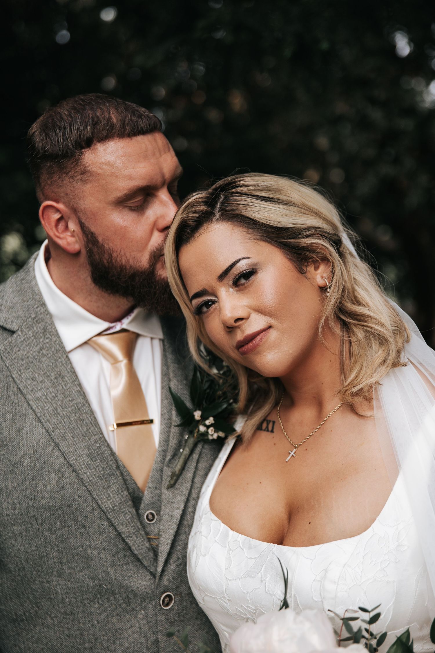 A bearded groom in a gray suit kisses his blonde bride in a white dress during their wedding portrait outdoors.