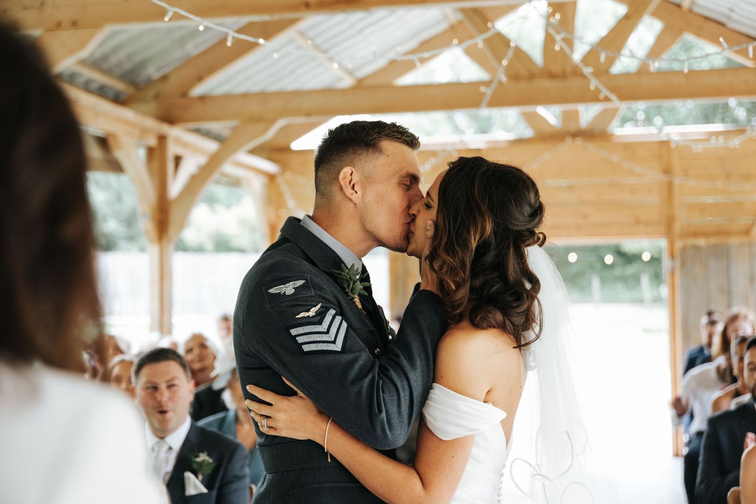 Military groom in uniform shares first kiss with bride in off-shoulder wedding dress at rustic barn ceremony.