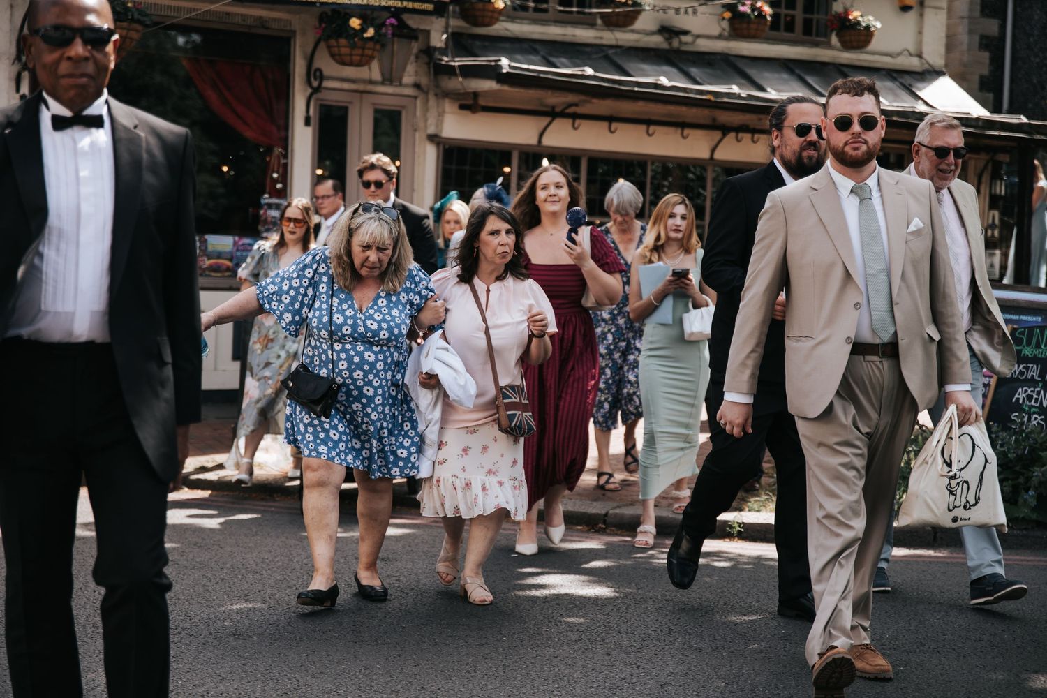 A group of elegantly dressed wedding guests walking down a sunny city street, wearing formal attire including floral dresses and suits.