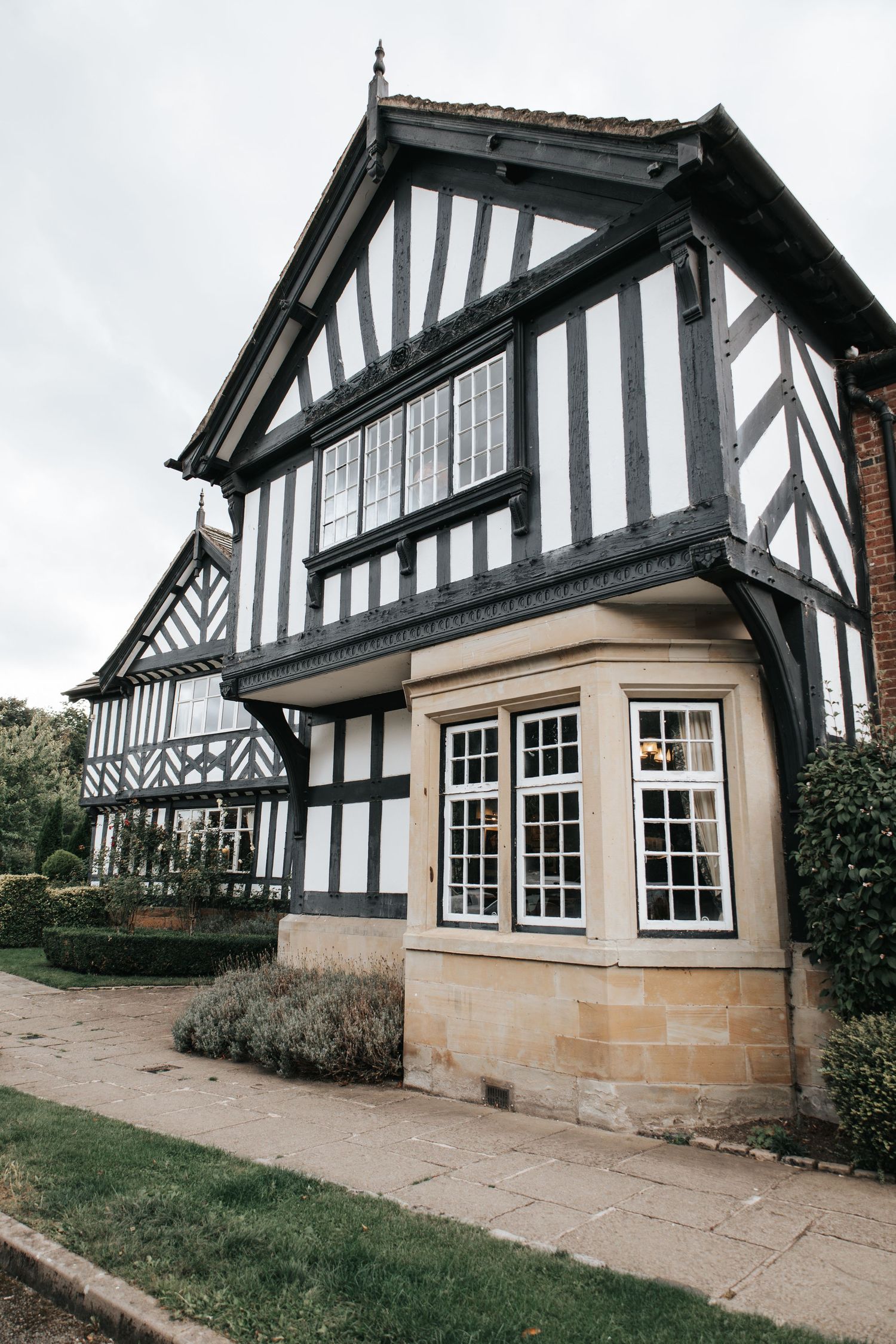 Historic Tudor-style building with black and white timber framing and stone bay windows surrounded by manicured gardens.