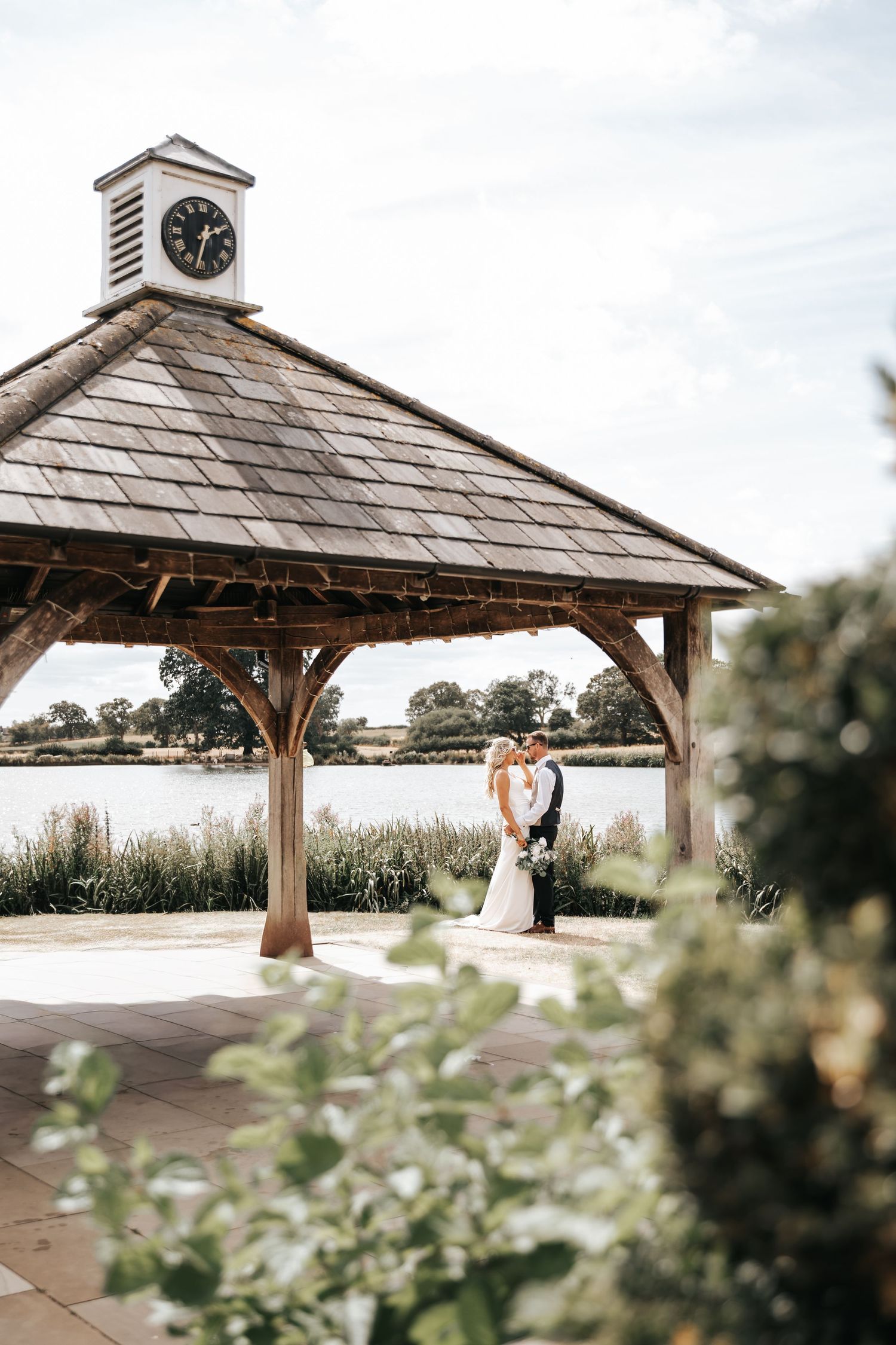 Bride and groom kissing under a rustic wooden gazebo with clock tower beside a serene lake on their wedding day.