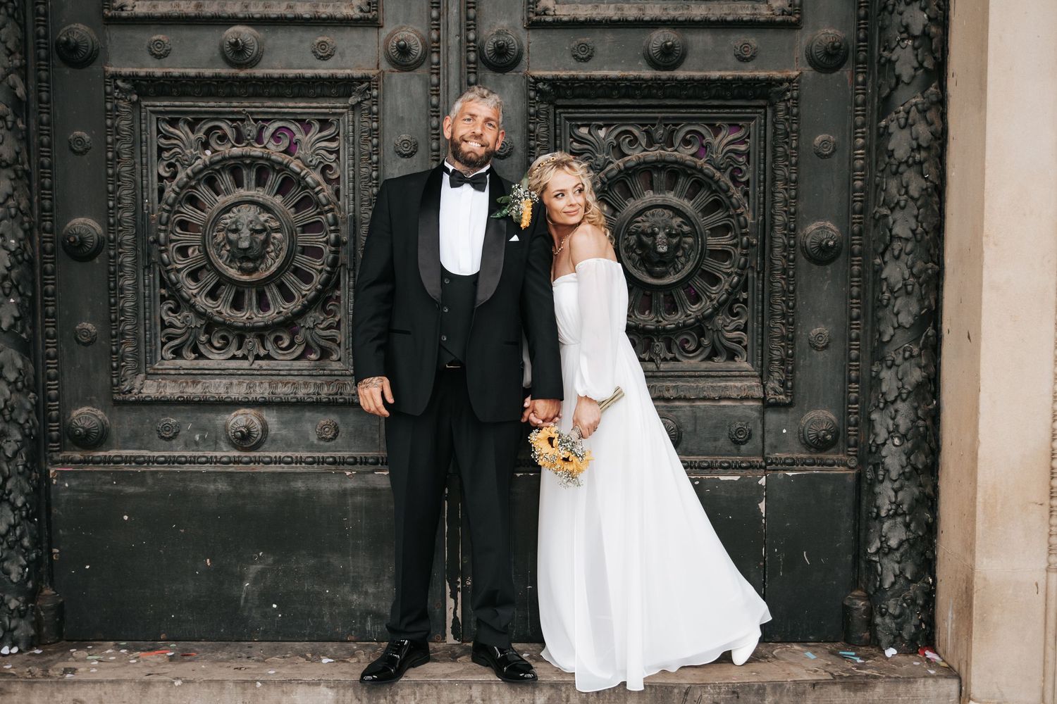 Bride and groom posing in wedding attire before ornate dark doors, man in tuxedo, woman in white gown holding bouquet.