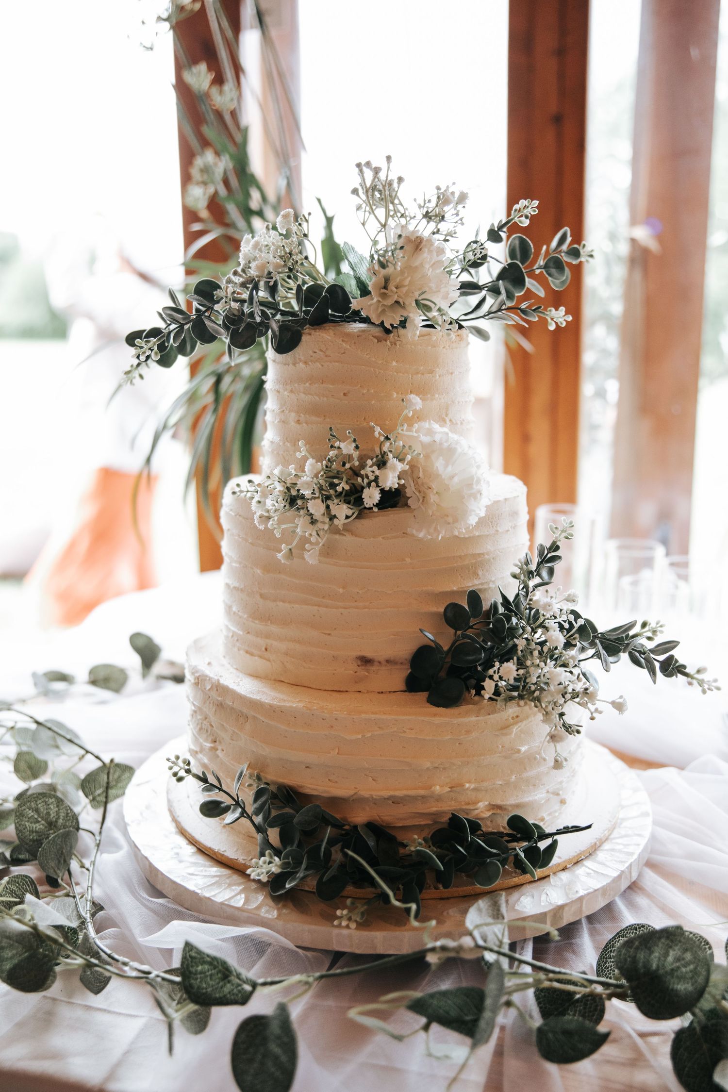 Three-tier white buttercream wedding cake adorned with eucalyptus and baby's breath florals on a draped table.