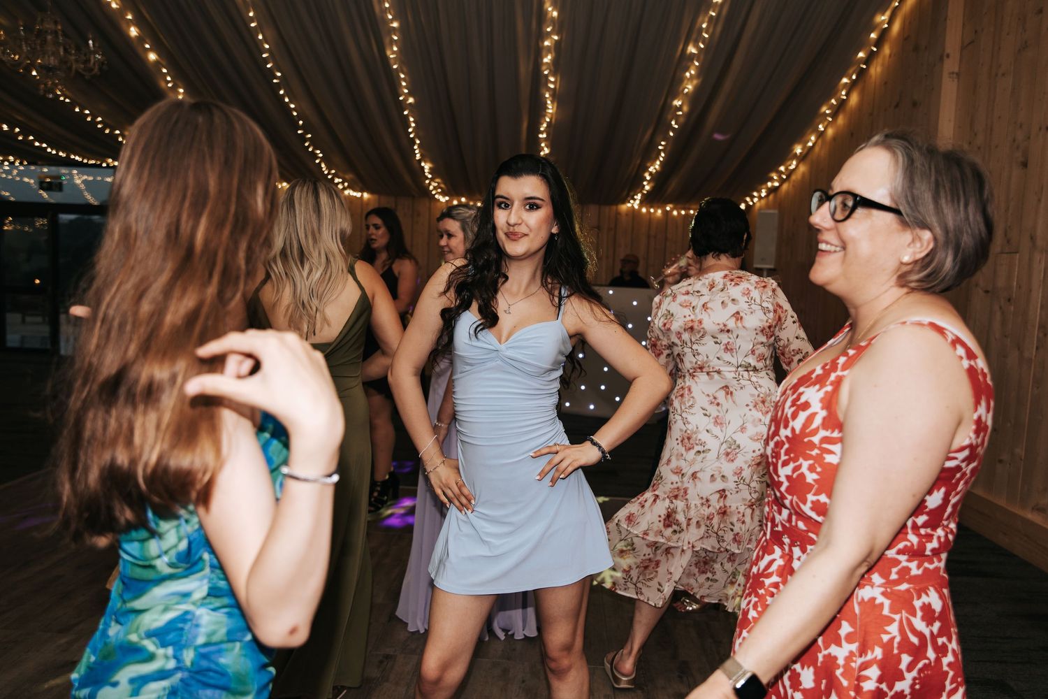 Women in cocktail dresses socializing and dancing at a fairy light adorned wedding reception.