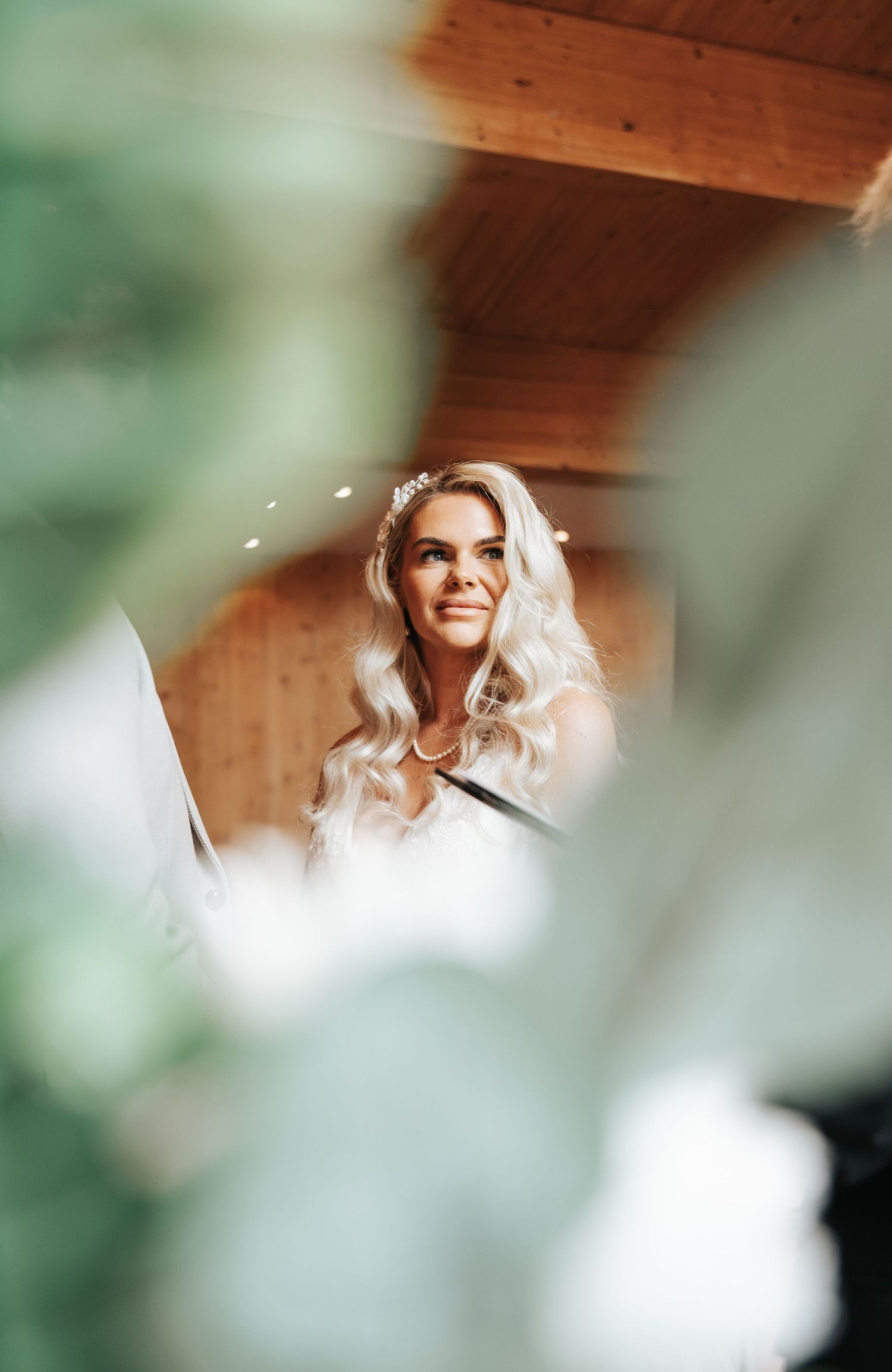 Smiling blonde bride with floral hair accessory in a wooden venue, captured through green foliage foreground.