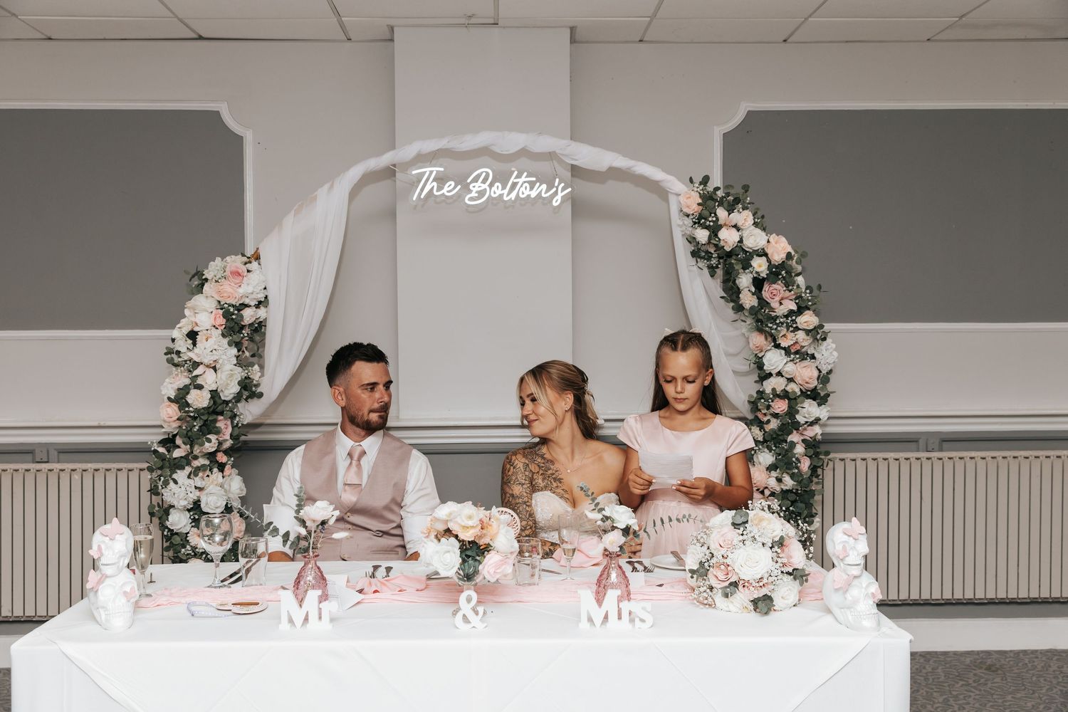 Bride and groom seated at elegant floral sweetheart table with pink roses and neon sign reading The Baltins.