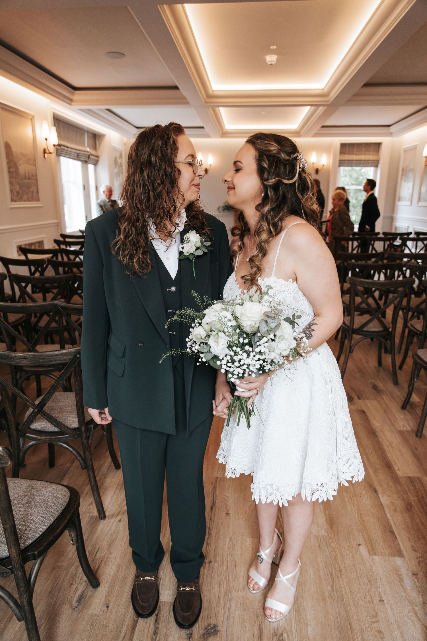 Two brides holding hands and gazing at each other during wedding ceremony, one in white lace dress, other in dark suit.