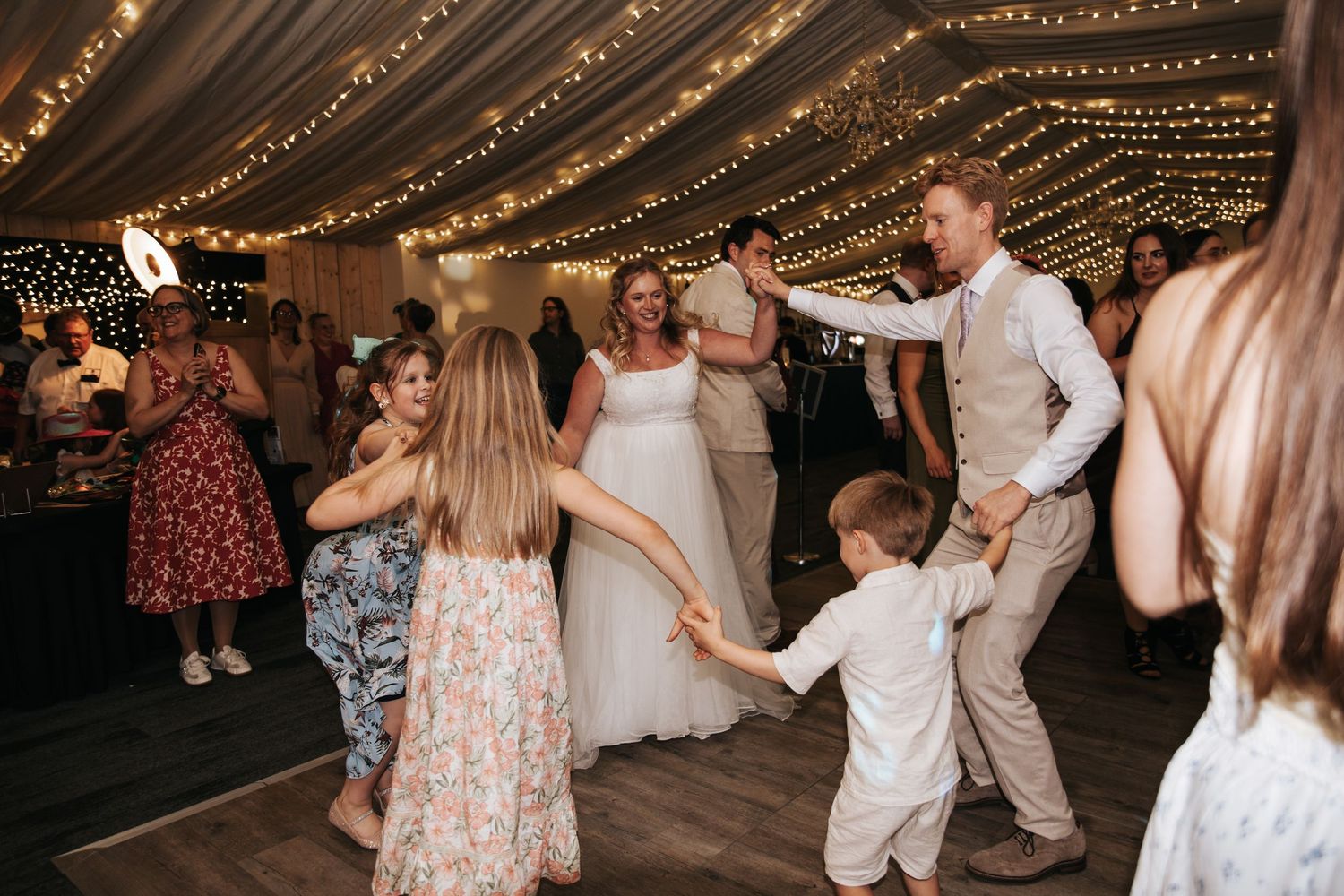 Bride and groom dancing with children and guests at a fairy light decorated wedding reception venue.