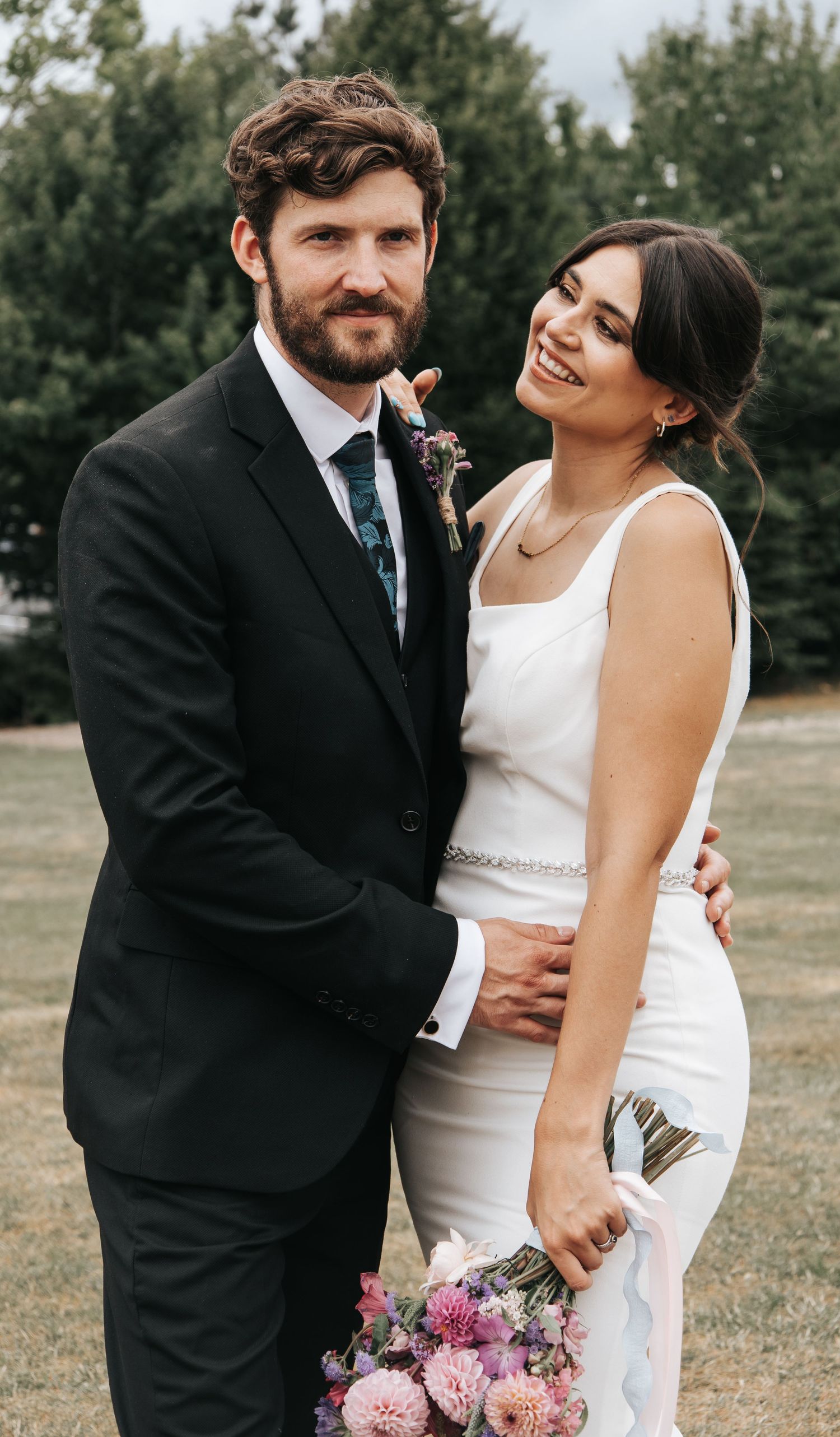 A joyful wedding couple embraces outdoors with natural greenery in the background.