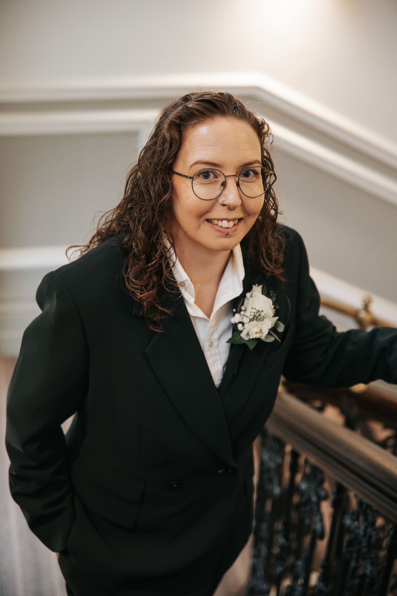Woman in black blazer with white boutonniere, glasses, curly hair, smiling on elegant staircase.