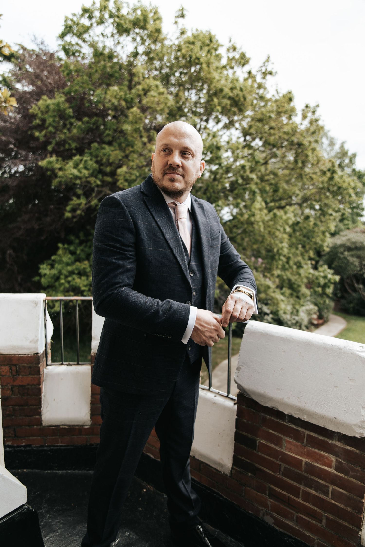 Bald man in dark suit with tie poses on outdoor terrace with brick walls and green trees behind.