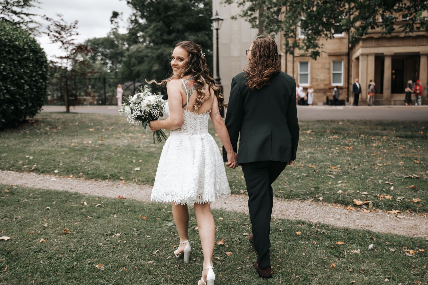 Bride in white lace dress holding bouquet walks hand-in-hand with groom in black suit on garden path.