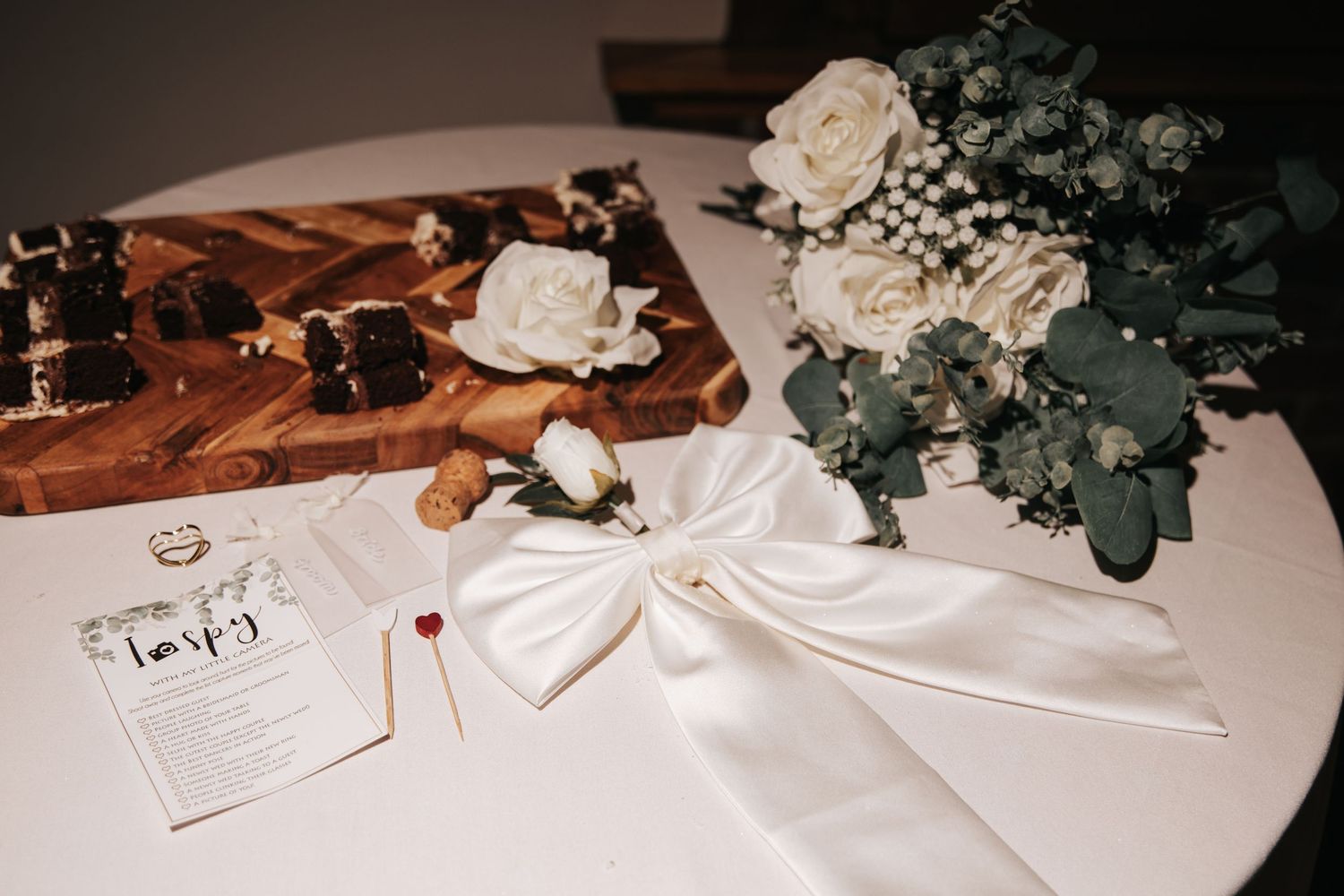 White wedding bouquet with roses and eucalyptus beside a satin bow, chess board, and elegant place card on a table.