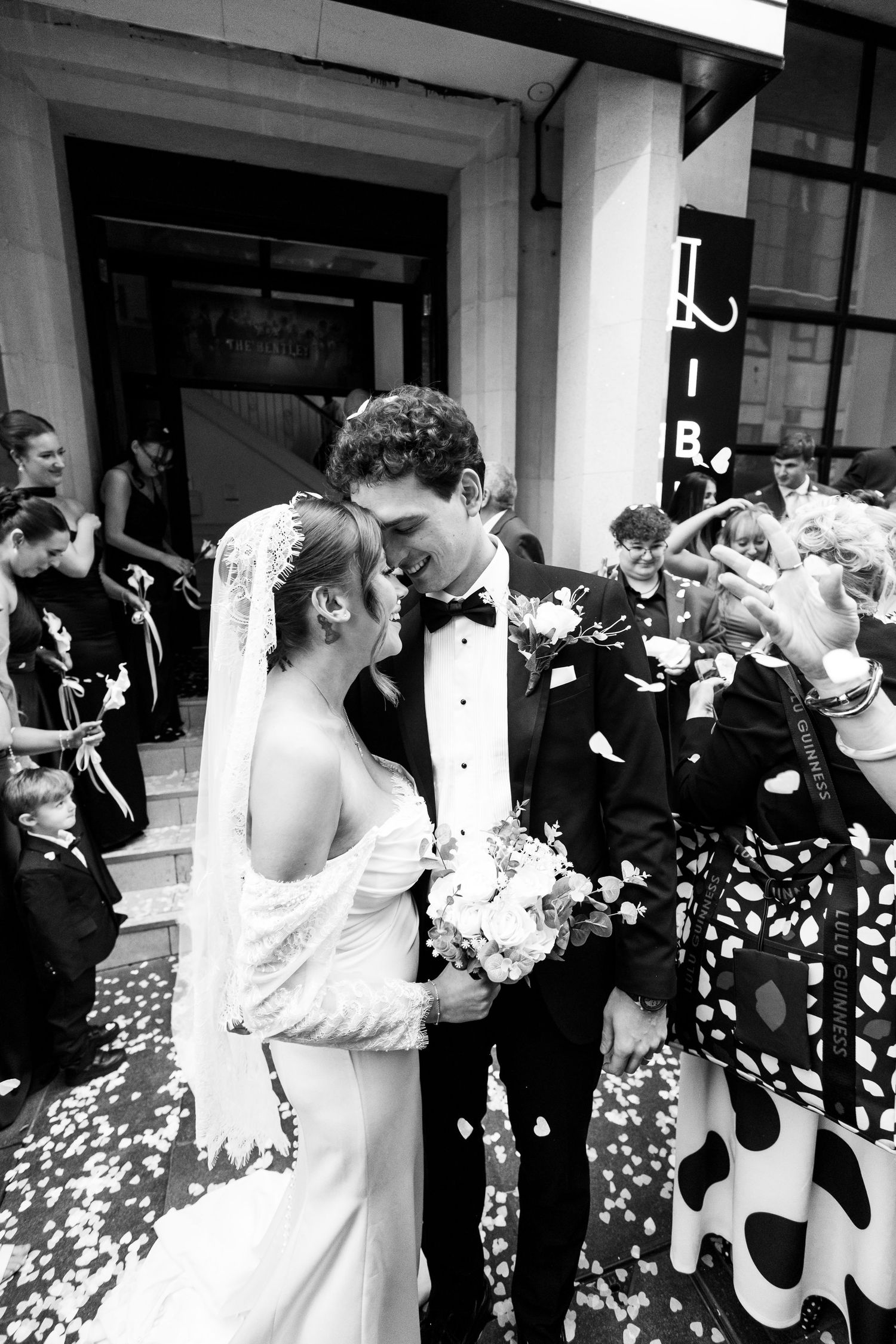 Black and white photo of a bride and groom sharing a romantic moment outside a church as guests throw confetti.