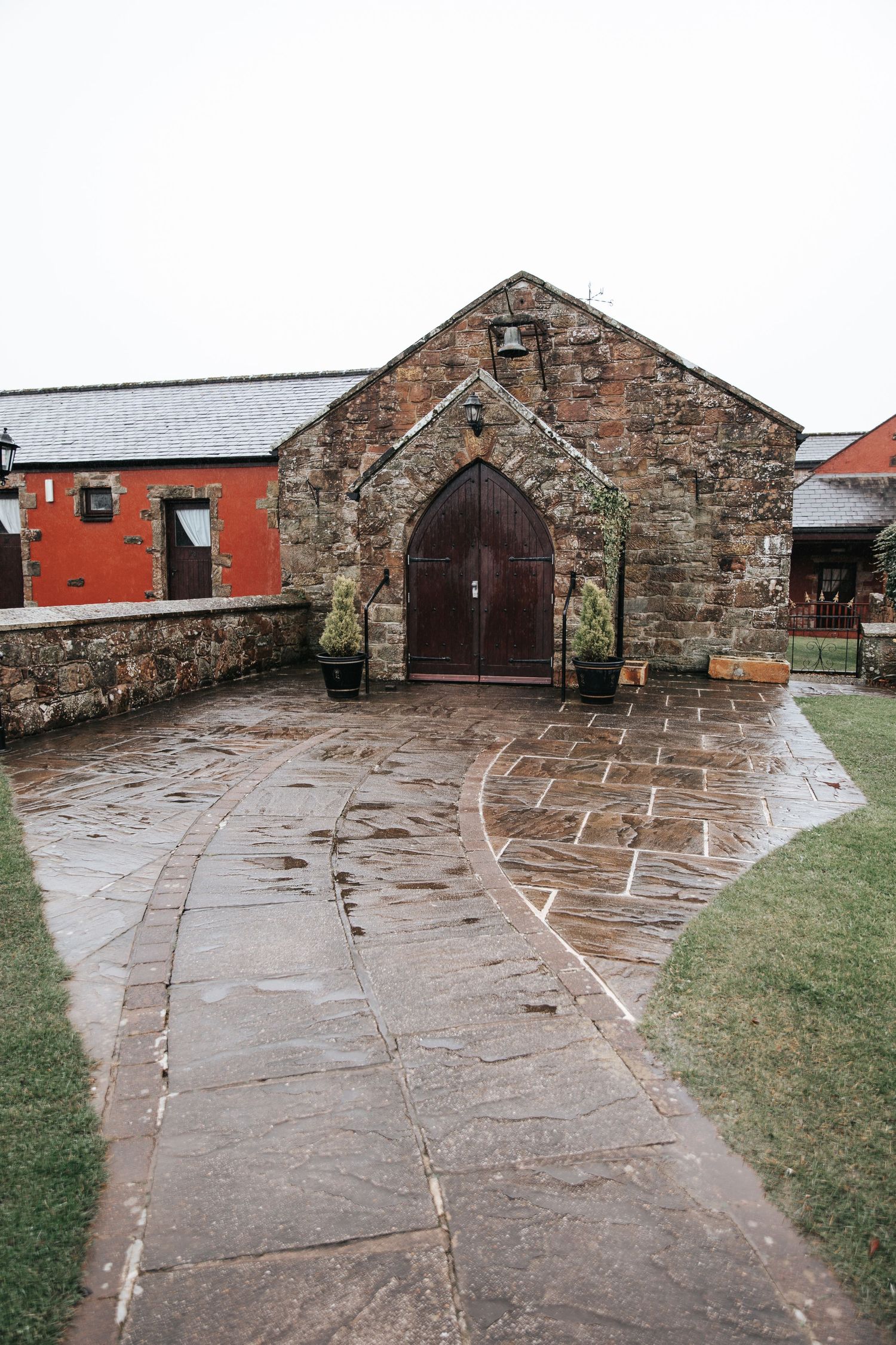Wet stone courtyard leads to a rustic chapel with wooden arched doors, flanked by red buildings on a rainy day.