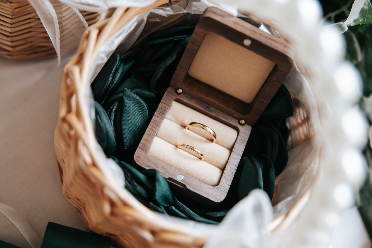 Two gold wedding bands displayed in a wooden ring box on green satin, nestled in a wicker basket.