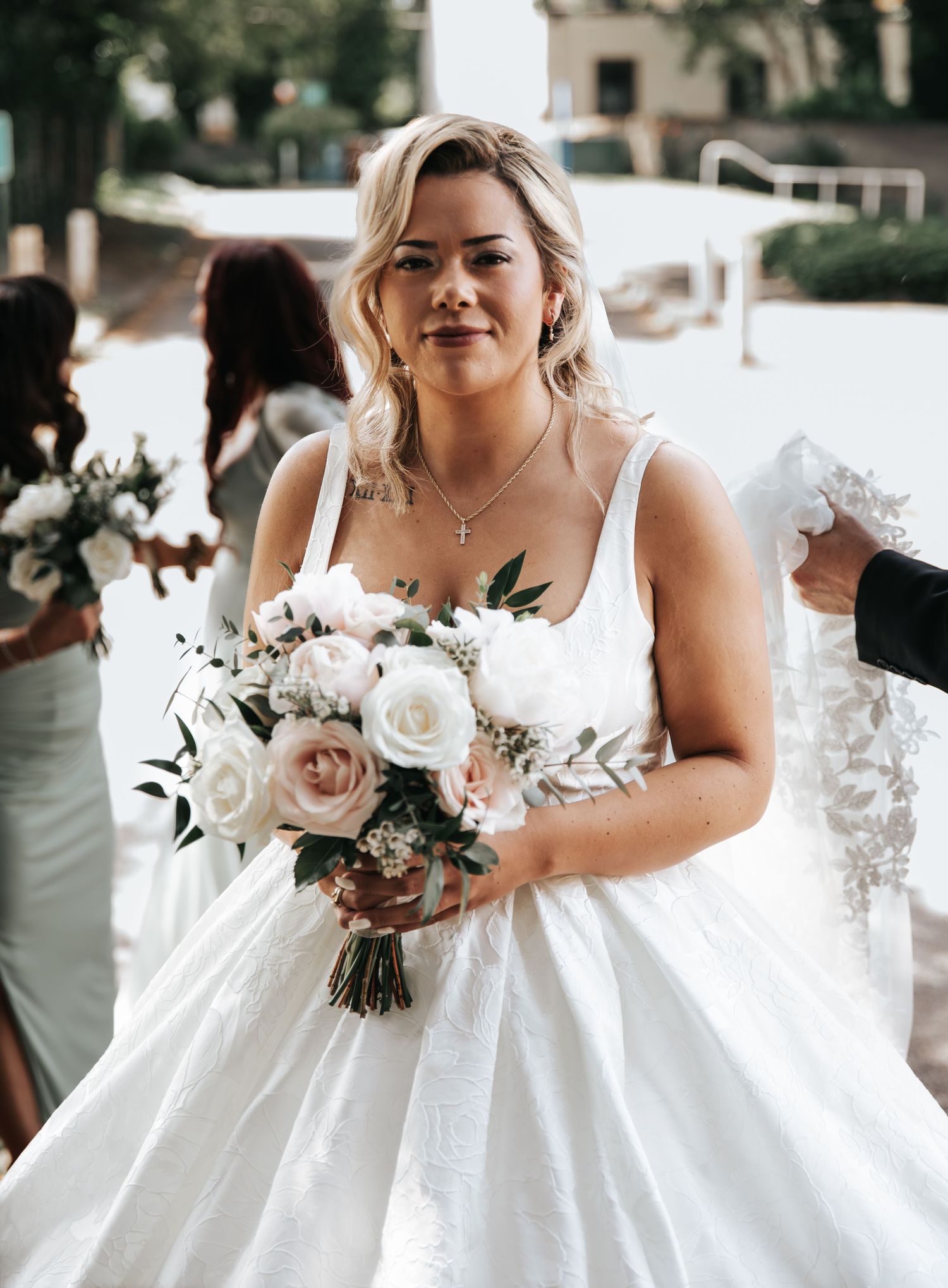 Smiling bride in white ball gown holding blush and white rose bouquet outdoors on her wedding day.