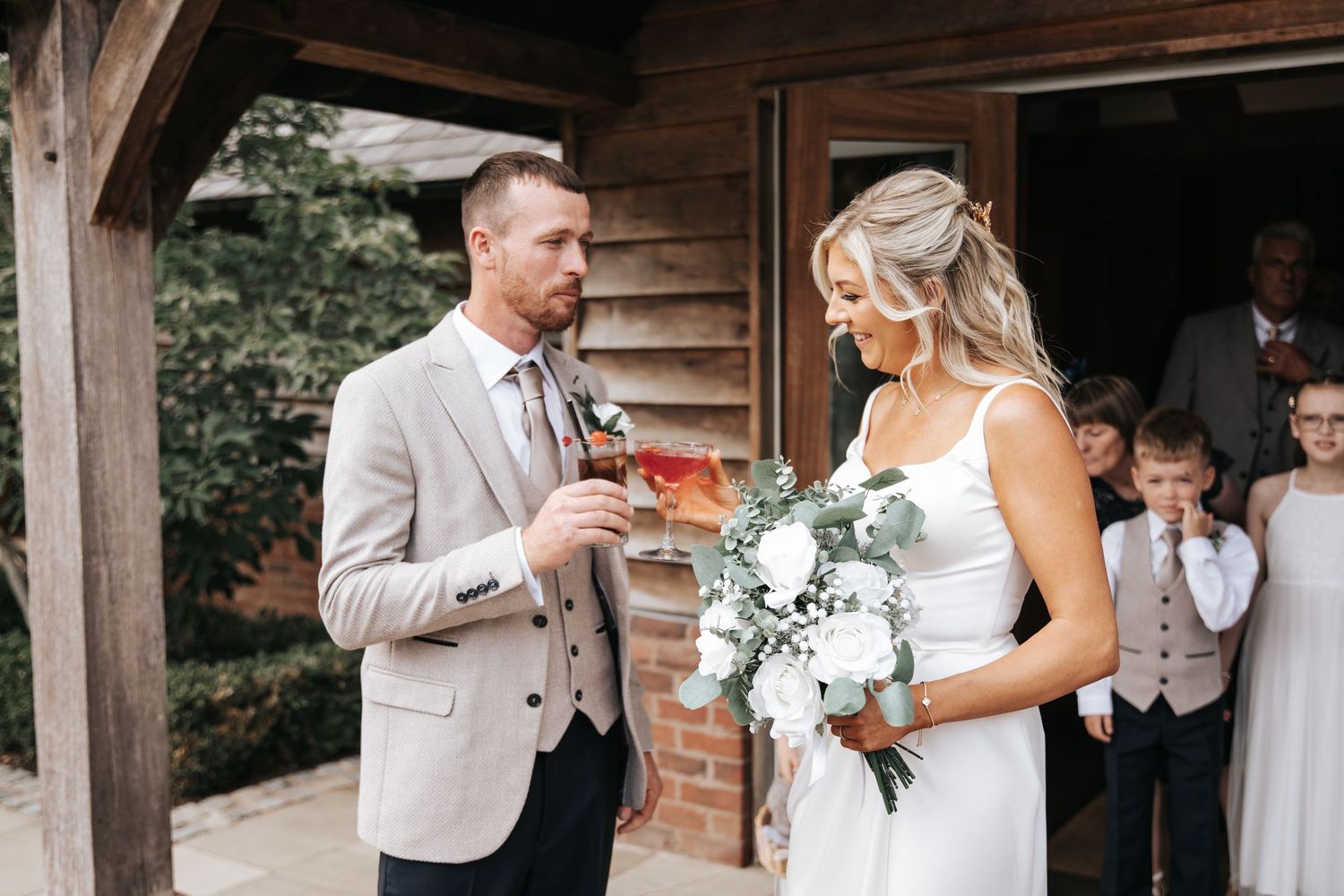 Bride holding white bouquet and groom in beige suit share a moment outside rustic wedding venue.