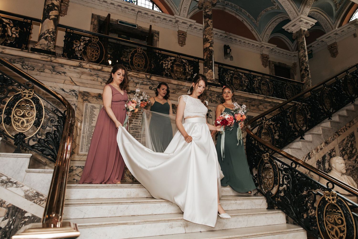 Bride in white gown with bridesmaids in colorful dresses on ornate marble staircase holding bouquets.
