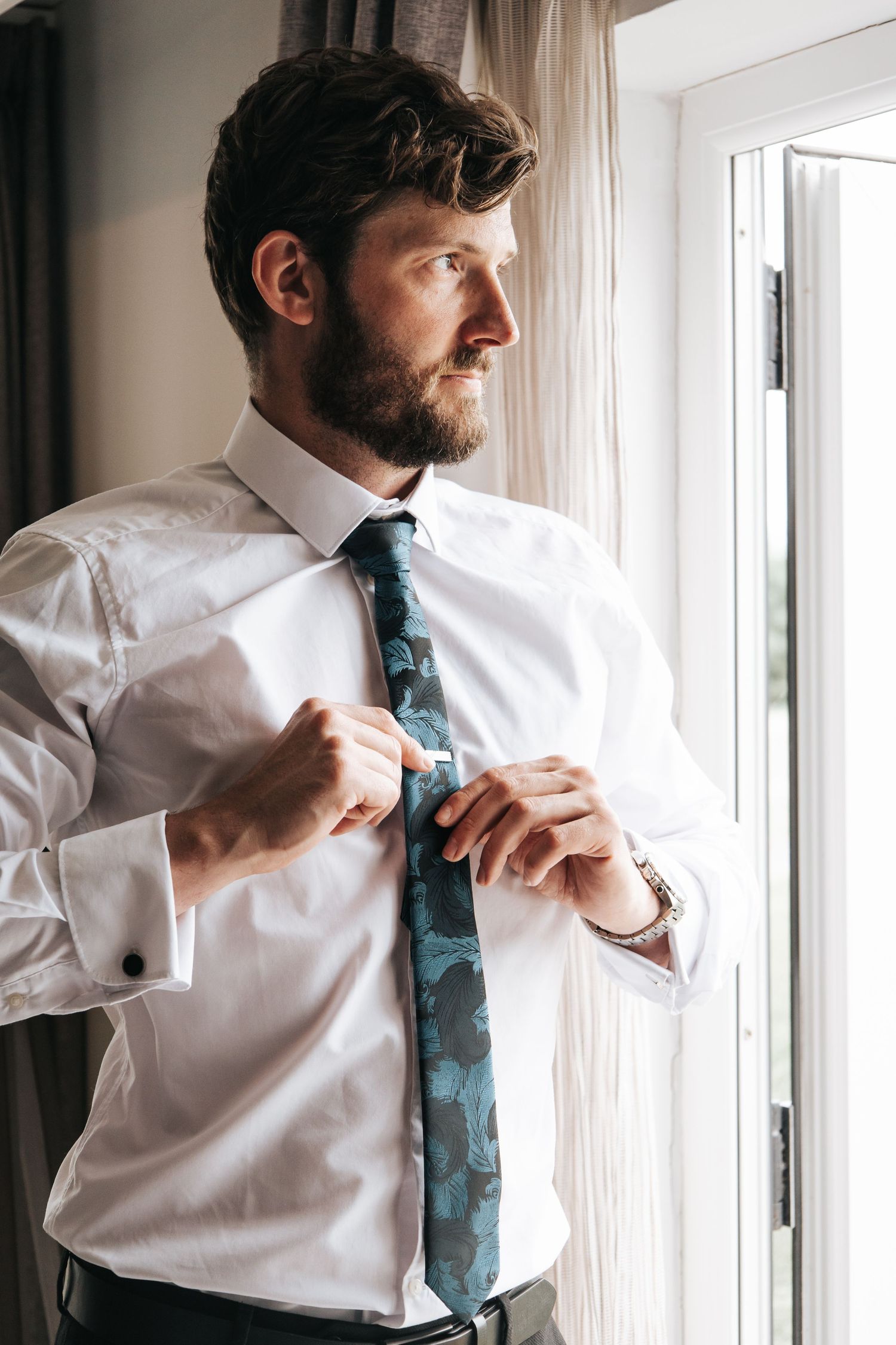 Man in white dress shirt adjusting blue patterned tie near window.