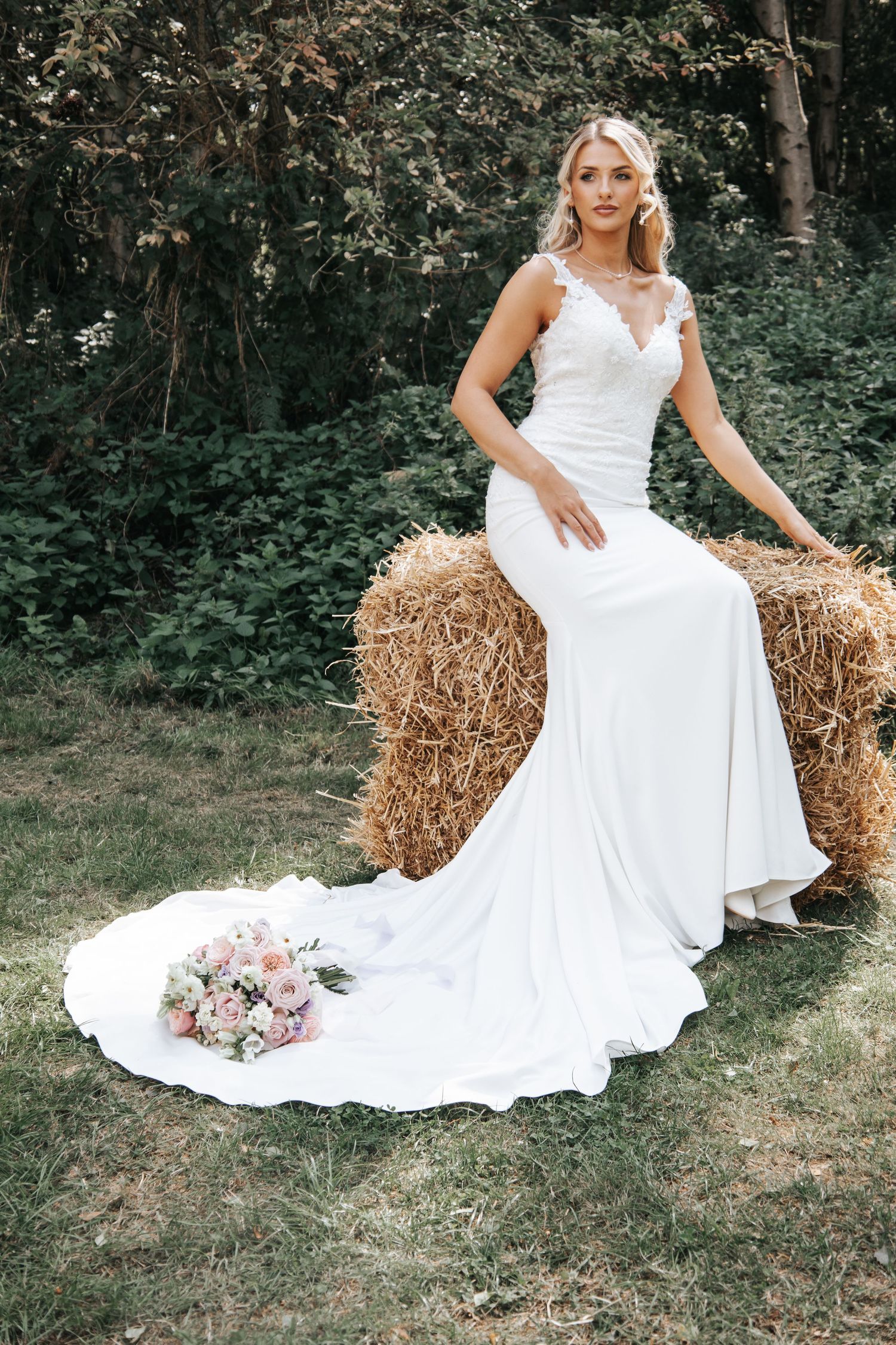 Wedding dress with flowing train displayed against hay bales with pink bouquet in outdoor garden setting.