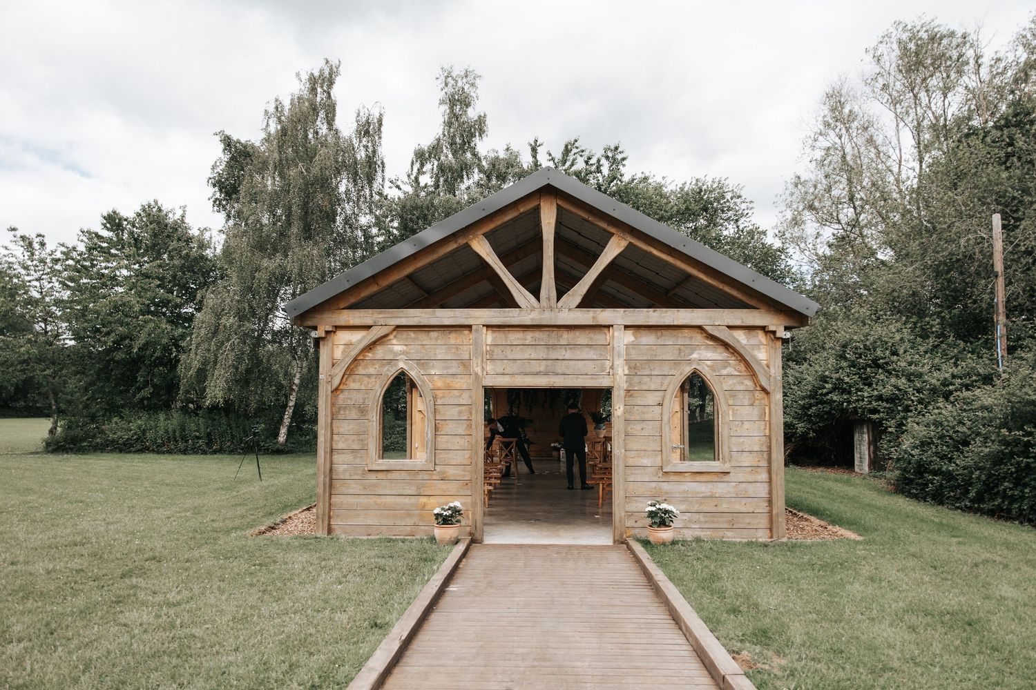 Rustic wooden chapel wedding venue with open doors, arched windows, and a gabled roof set in lush green countryside.