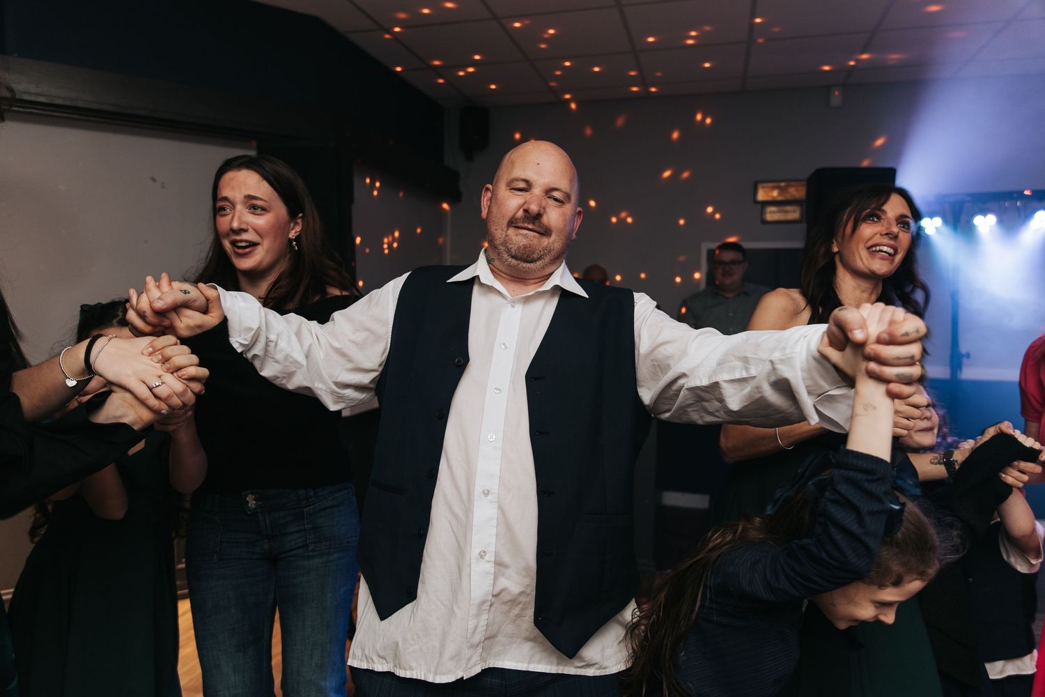 Wedding guests dance and celebrate on a dimly lit dance floor with red accent lighting above.