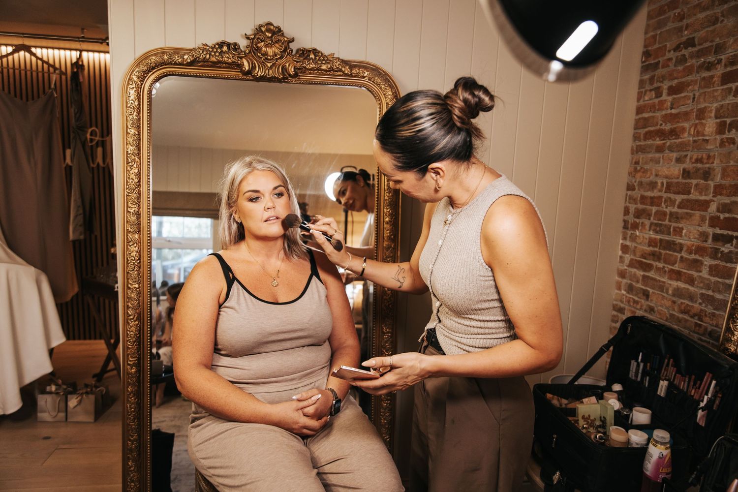 A makeup artist applies foundation to a seated woman in a beauty salon with a large ornate mirror in the background.