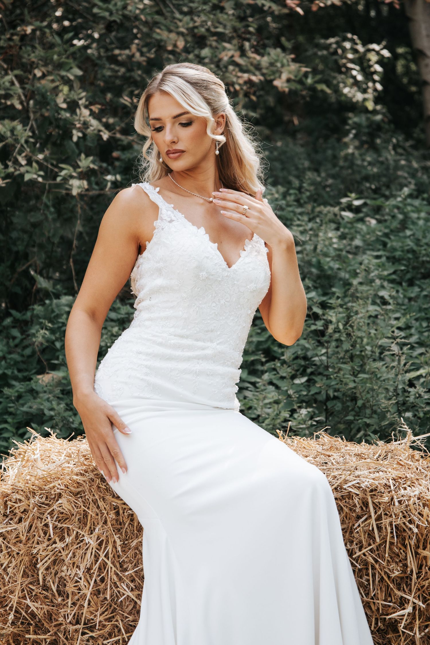 Bride wearing white lace v-neck wedding gown sits elegantly on hay bale with natural forest backdrop.