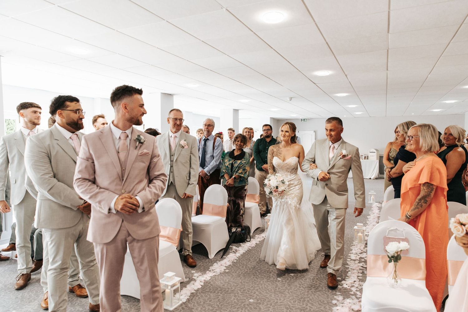Bride walks down the aisle in a white gown as groom smiles in a blush suit during an indoor wedding ceremony.