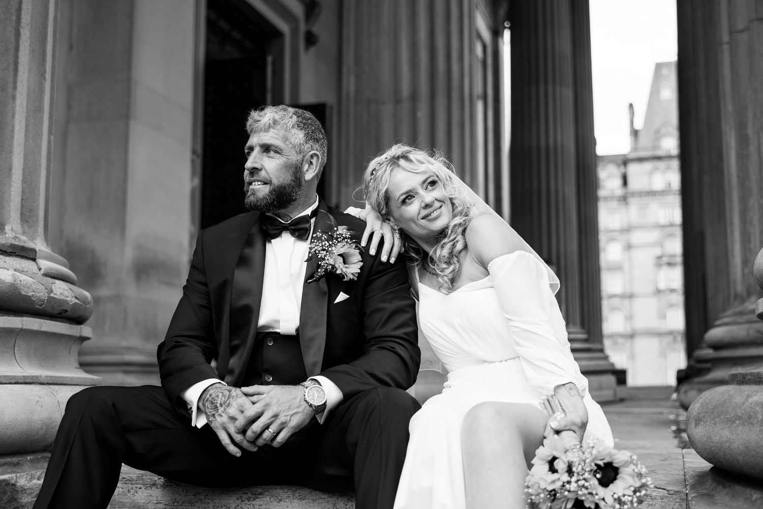 Black and white photo of a bride and groom sitting on steps, smiling, with columns in the background.
