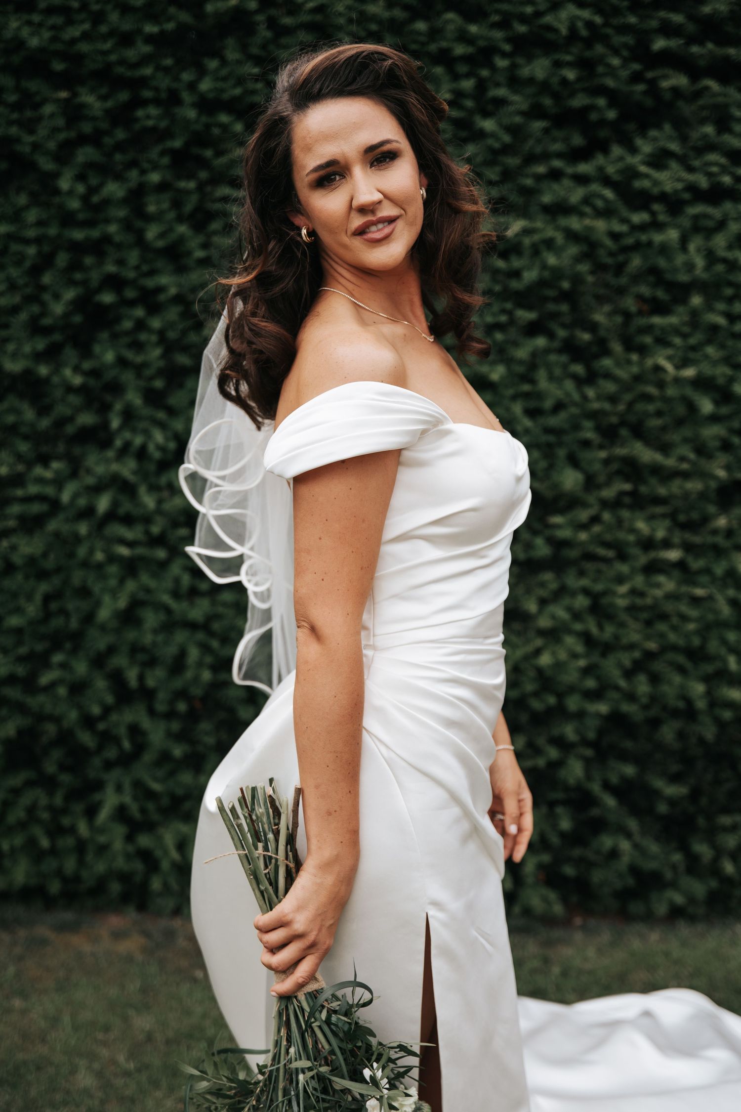 Bride in off-shoulder white gown holding greenery bouquet, posing before ivy wall.