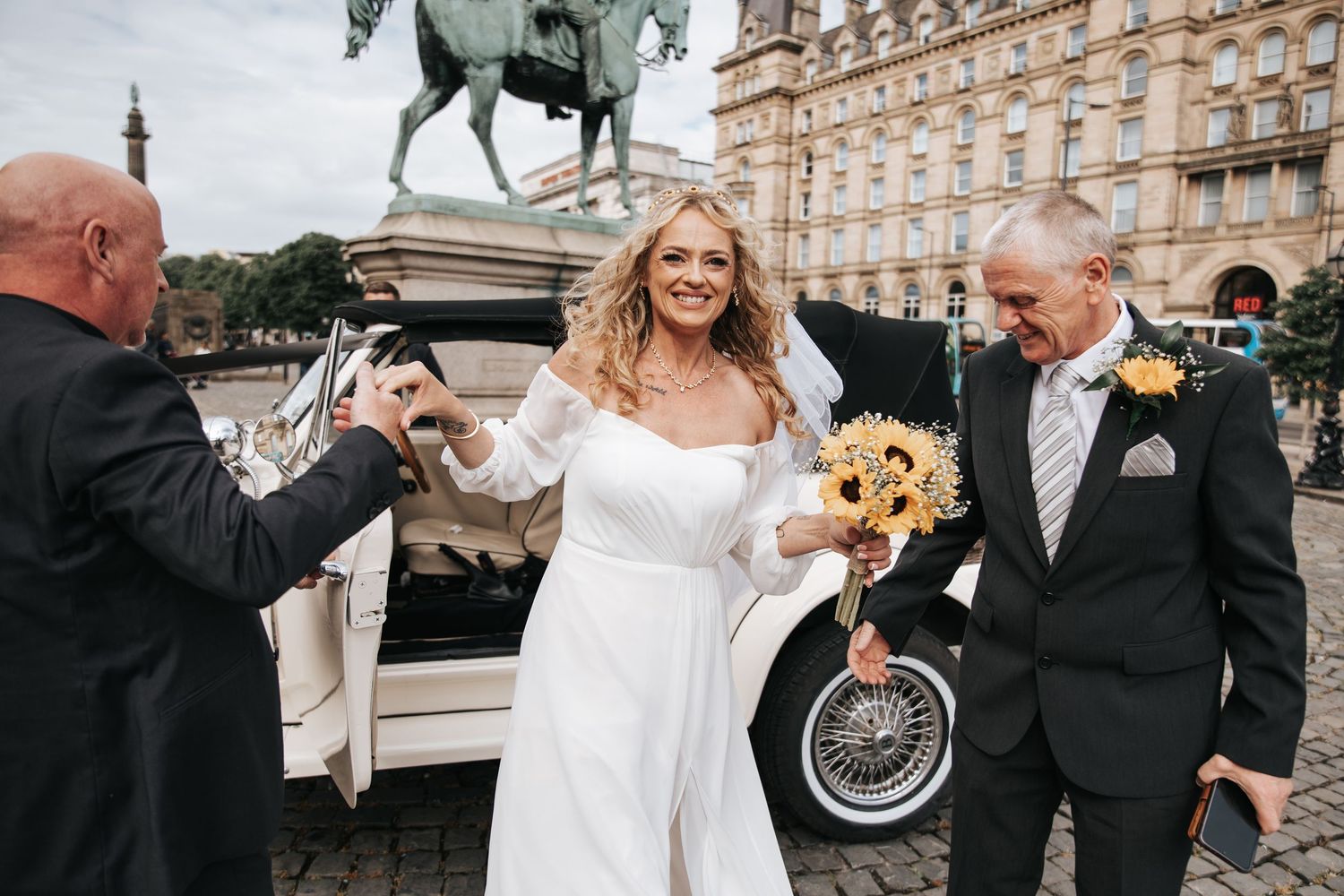 Bride in white gown holding sunflower bouquet exits vintage car with groom at outdoor wedding ceremony.
