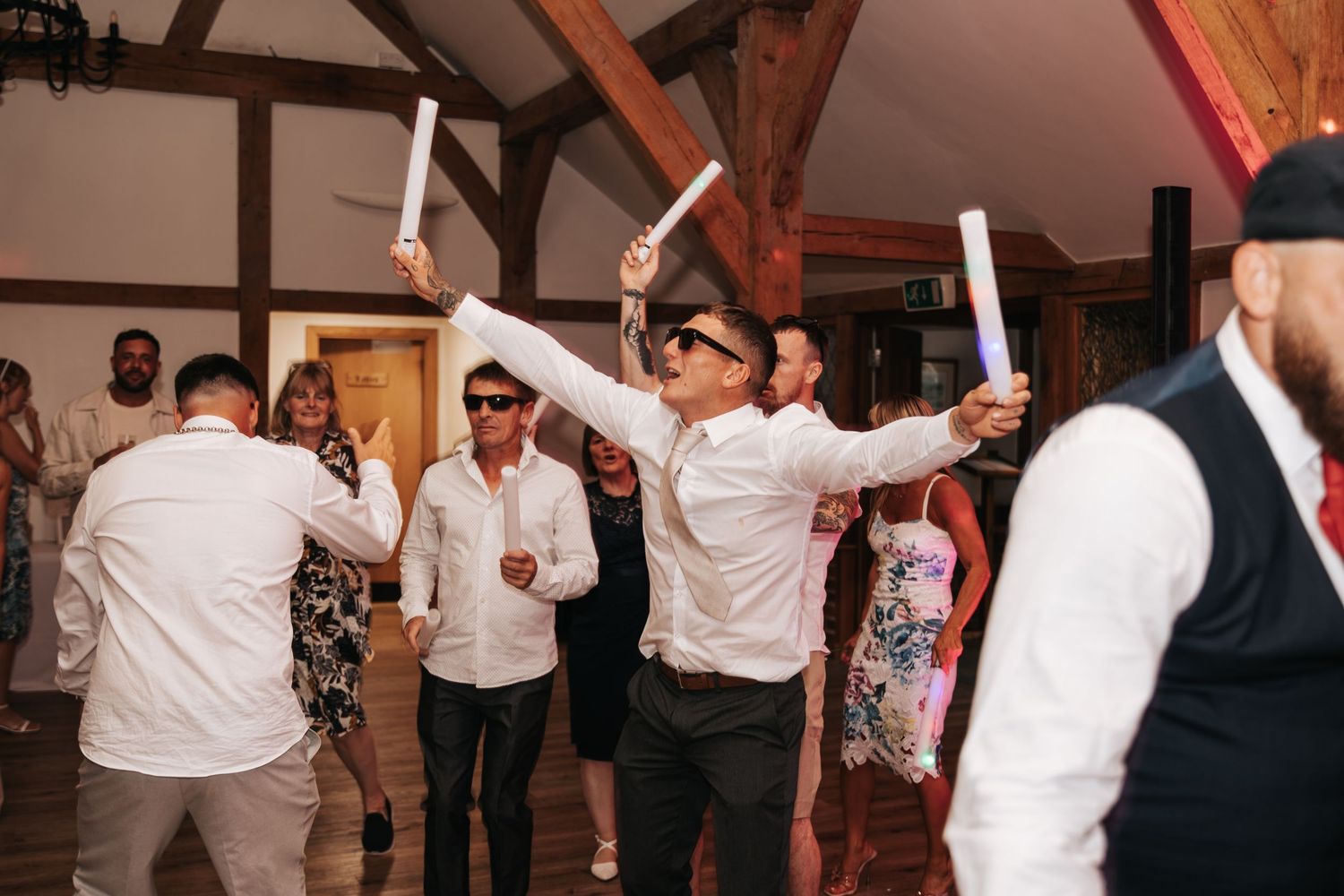 Guests dancing and waving glow sticks at a lively wedding reception in a rustic venue.
