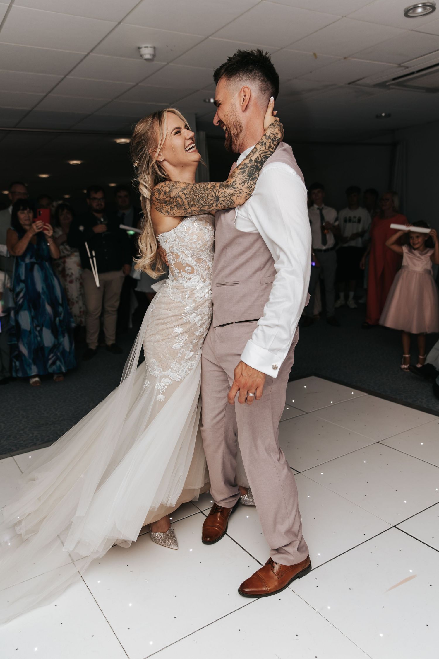 Bride and groom share their first dance, she in a lace gown, he in a grey suit with tattoos visible.