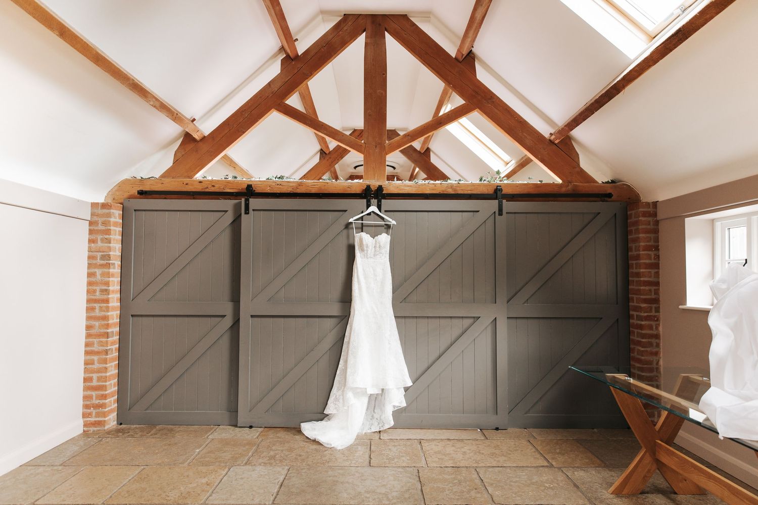 White wedding dress hanging on barn doors with exposed wooden beams and brick walls in a rustic venue.
