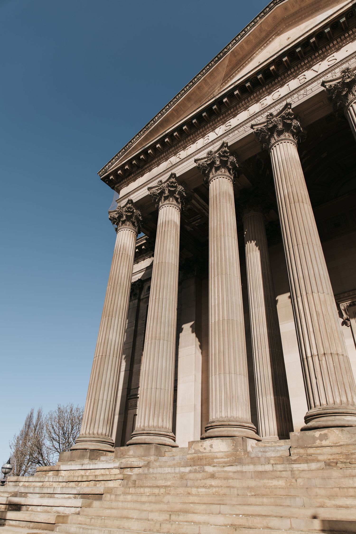 Neoclassical building facade with tall Corinthian columns, stone steps, and ornate cornice against a clear blue sky.
