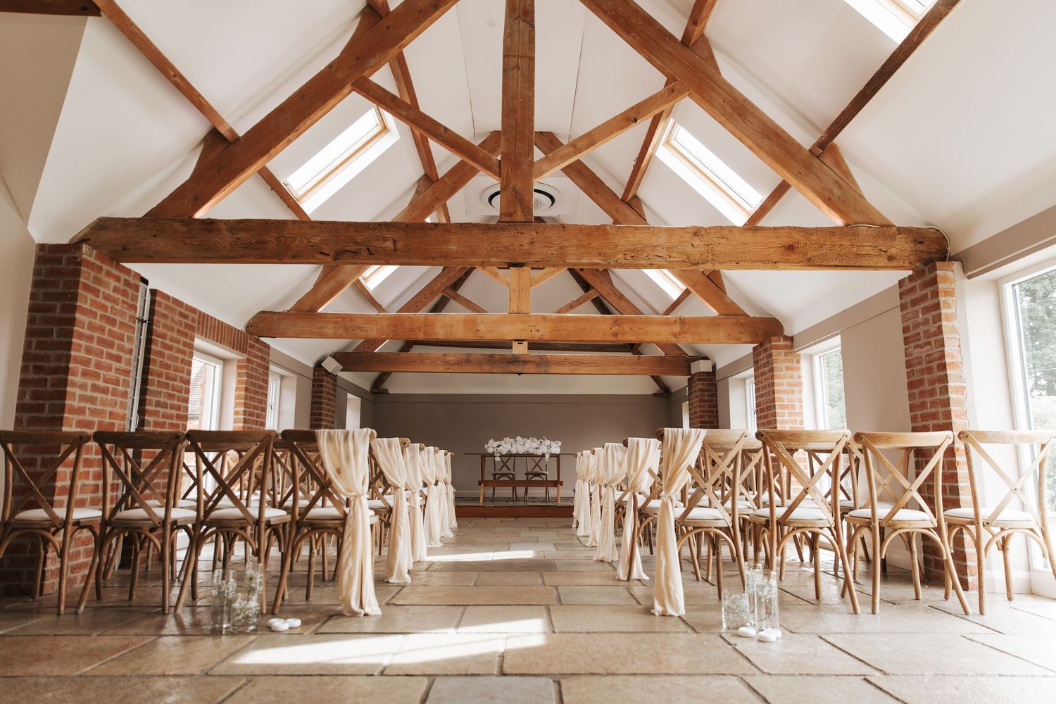 Elegant barn wedding venue with wooden beamed ceiling, cross-back chairs, and stone floor aisle.