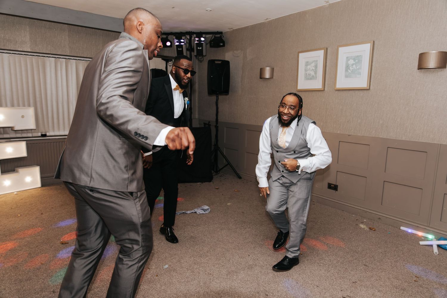 People in formal attire dance and celebrate at an indoor party with speakers and wall art visible in the background.