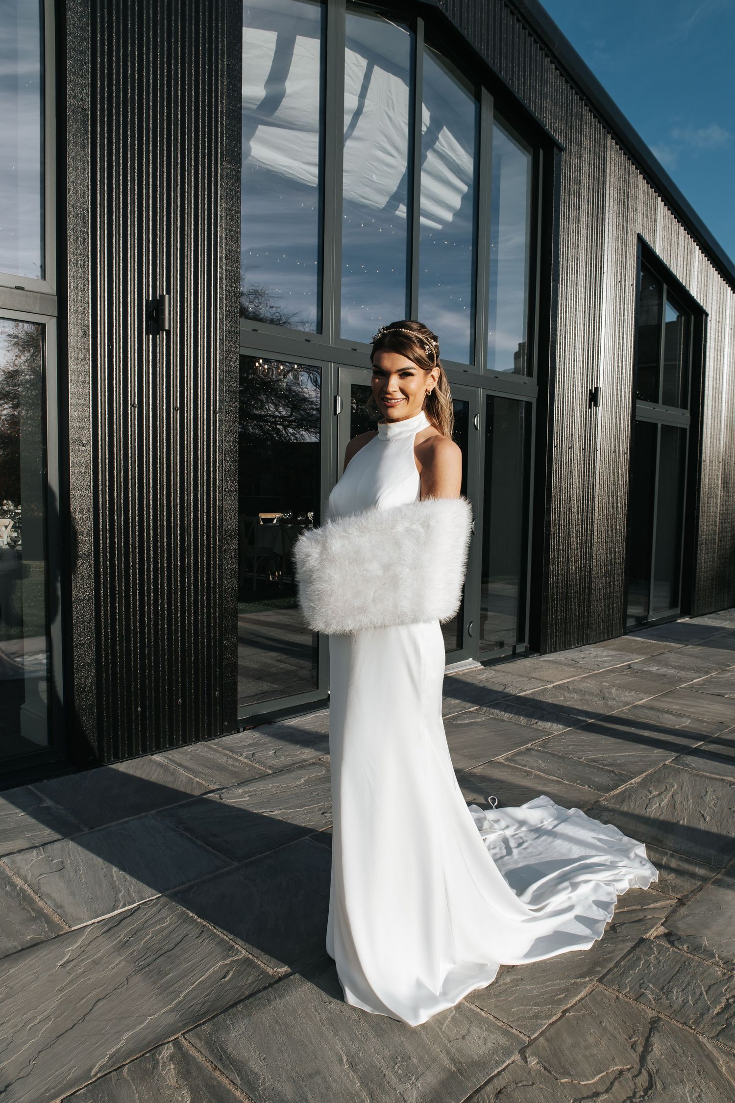 Modern bride in sleek white dress and fur wrap standing against industrial building exterior.