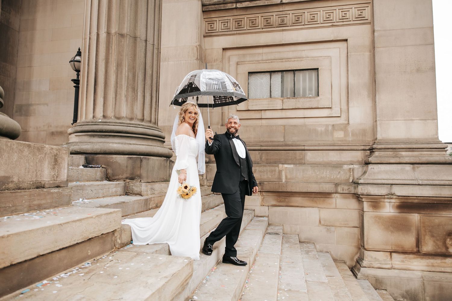 Bride and groom walking down stone steps under a clear umbrella outside a grand classical building.