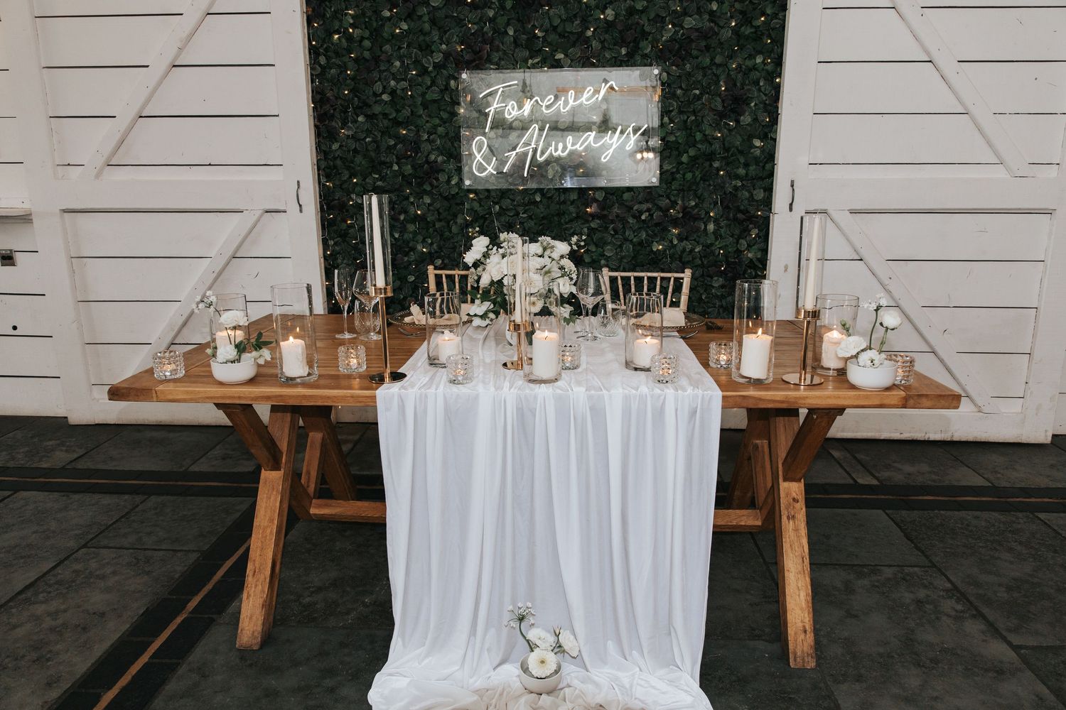 Rustic wooden sweetheart table with white chiffon runner, candles, and floral centerpiece before a green hedge wall.