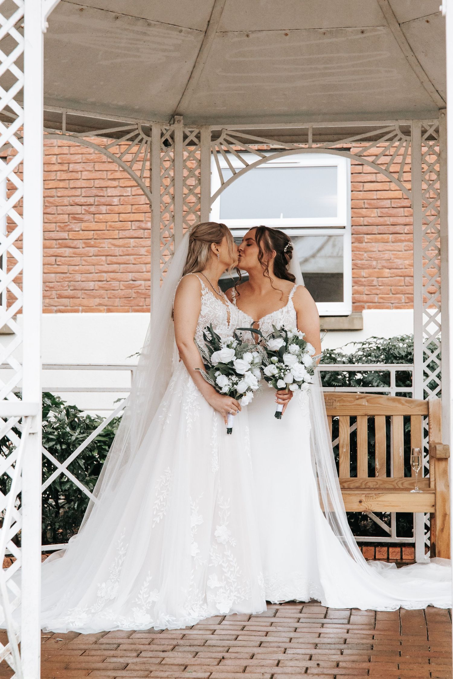 Two brides in white gowns pose under white gazebo with brick building in background.