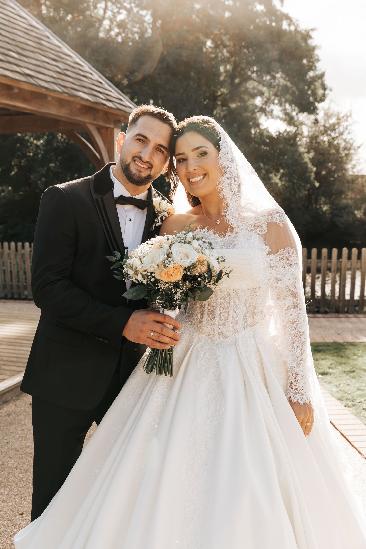 Smiling bride and groom pose outdoors in wedding attire, bride holding white and peach bouquet with lace veil.