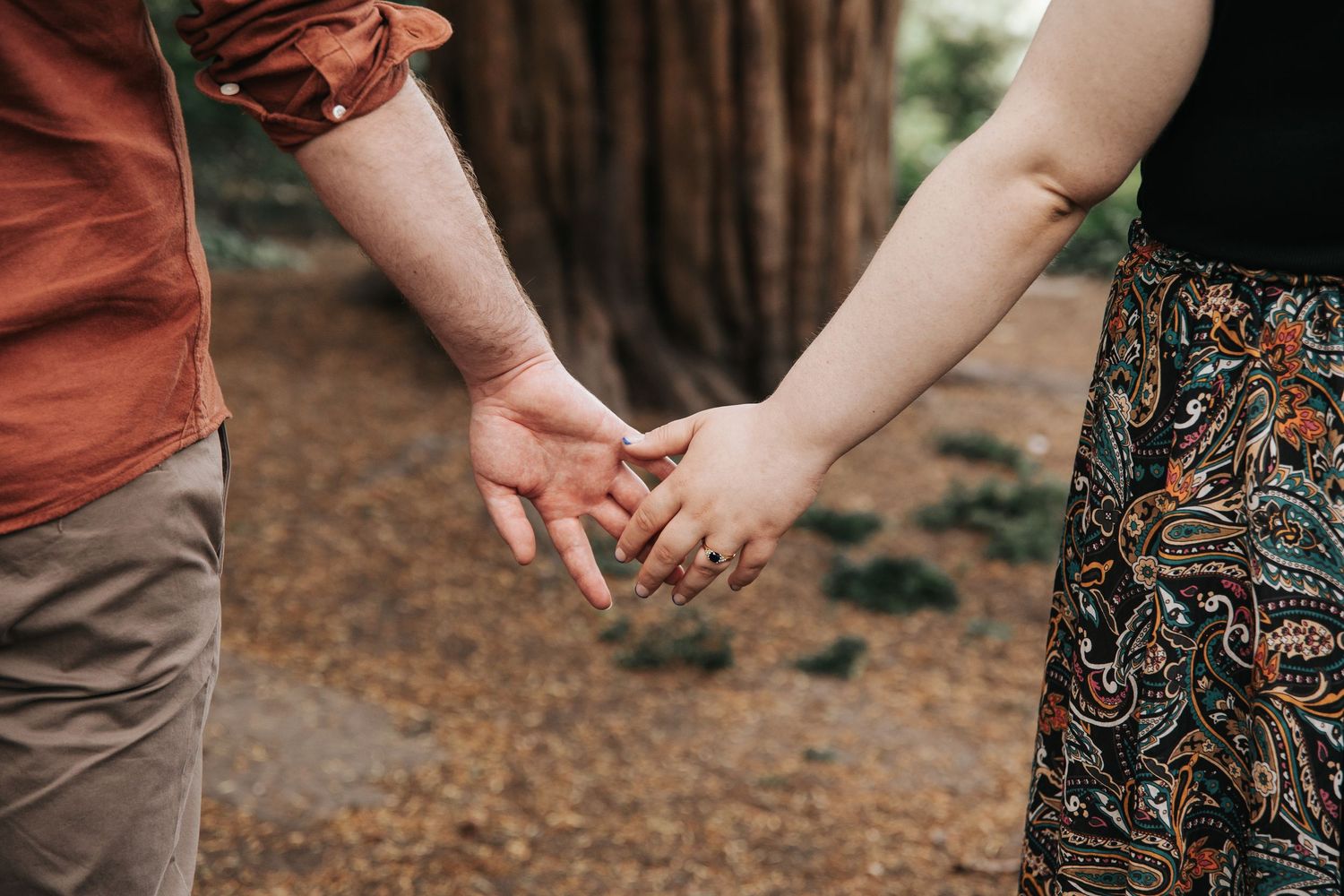 Couple holding hands outdoors in a forest setting, woman wearing a paisley skirt and ring.