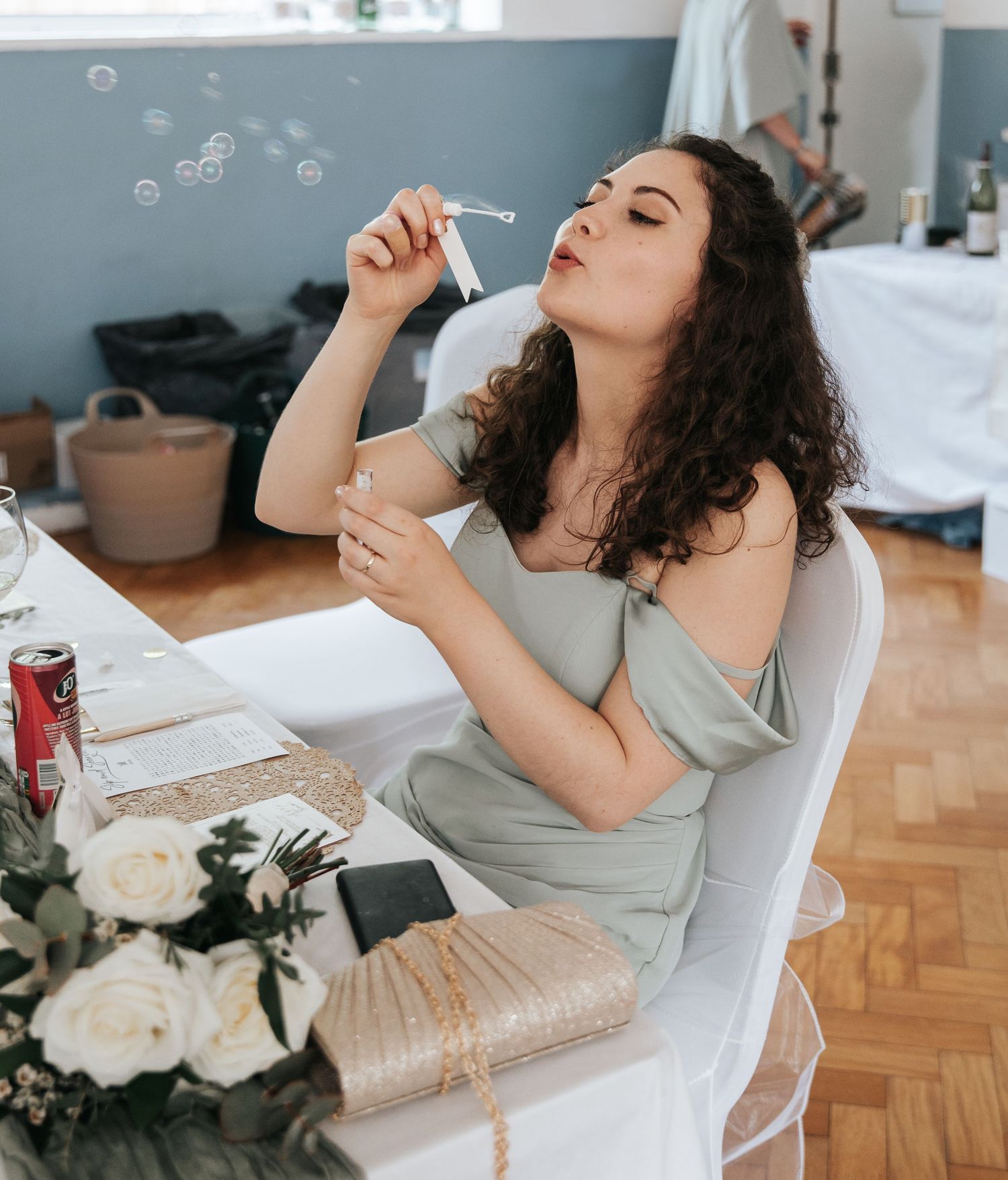 Woman in sage green dress blowing bubbles at a wedding reception, seated at a decorated table with white roses.