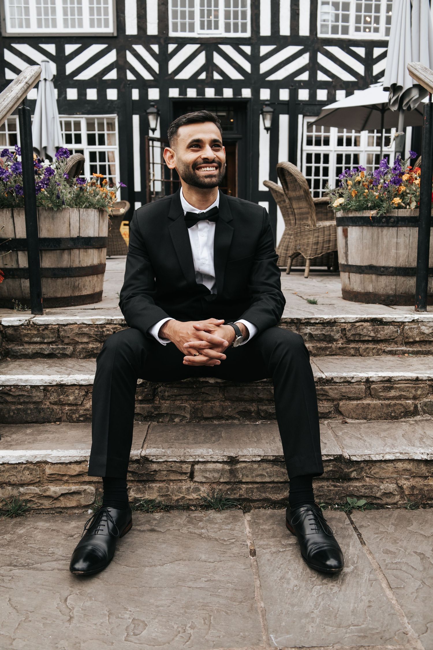 Groom in classic tuxedo sits casually on stone steps of Tudor-style building.